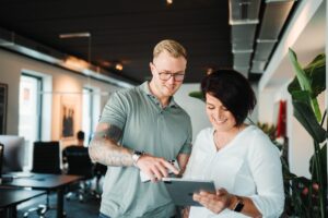 Zwei Personen, ein Mann und eine Frau, nutzen in einem modernen Büro gemeinsam ein Tablet. Sie arbeiten fokussiert an einer digitalen Aufgabe, um das Prinzip der Eisenhower Matrix zu verdeutlichen.