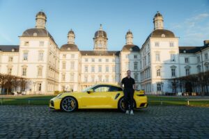 Ein Mann lehnt lässig an einem hochwertigen, gelben Sportwagen vor einem imposanten Schloss. Das Bild verdeutlicht die Bedeutung von Timeboxing für Entscheider: Es symbolisiert den Erfolg und die Freiheit, die durch gezielte Zeitblöcke für wichtige Aufgaben entstehen, um maximale Effizienz und beeindruckende Ergebnisse zu erzielen.