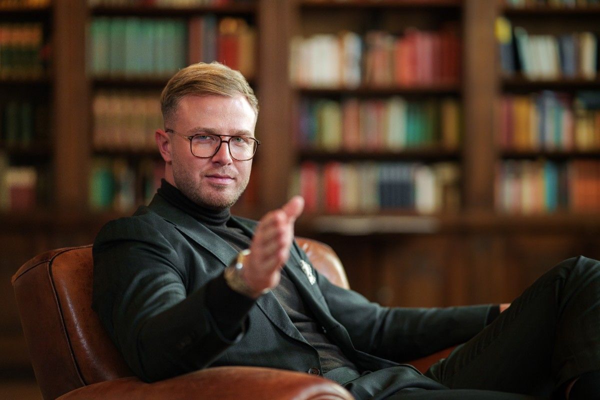 Ein selbstbewusst auftretender Mann mit Brille sitzt in einem Ledersessel vor einer Bücherwand und blickt direkt in die Kamera. Kompetenz, Erfolg und strategisches Denken werden durch die seriöse Kleidung und die gelehrte Umgebung symbolisiert.
