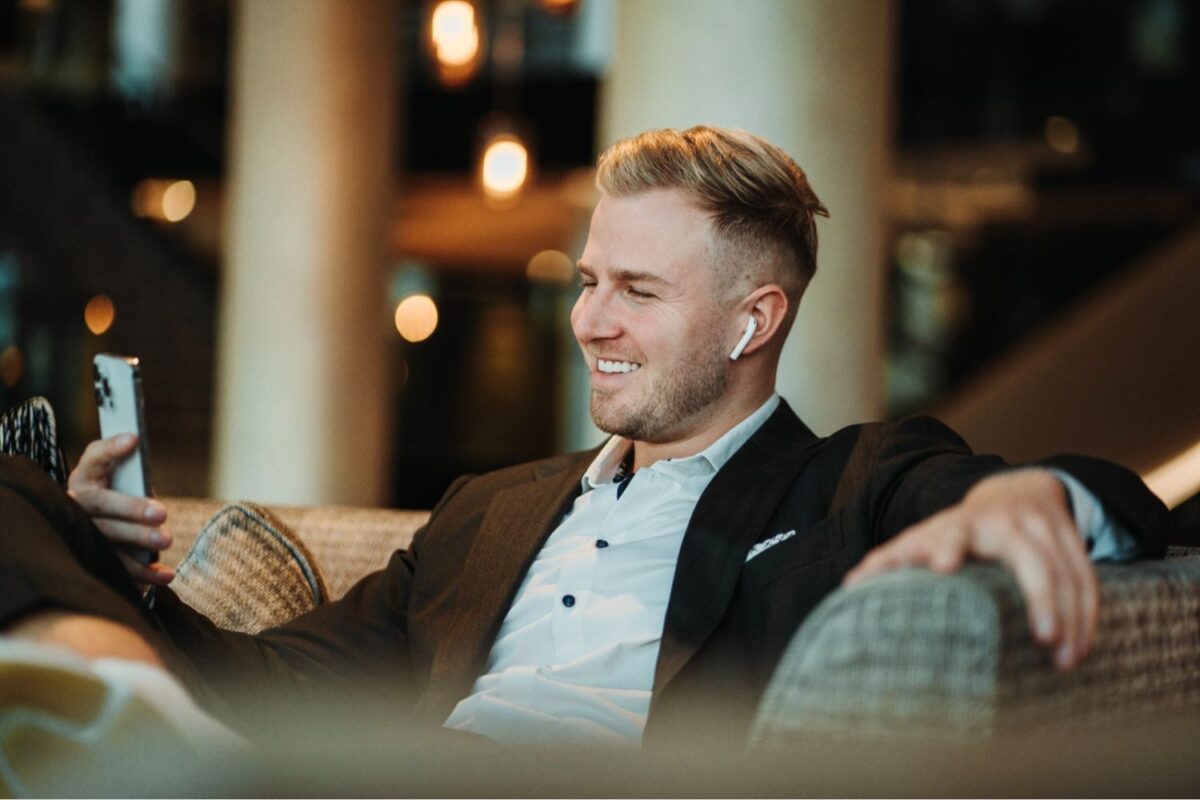 Entspannter Geschäftsmann sitzt lächelnd mit Smartphone auf dem Sofa, als Resultat von Persönlichkeitsentwicklung für die Umsatzsteigerung.