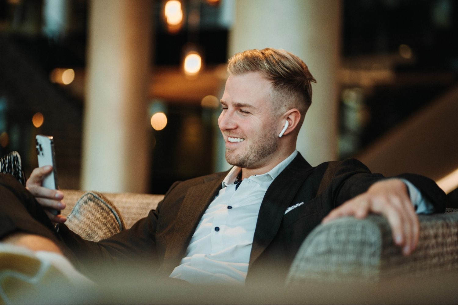 Entspannter Geschäftsmann sitzt lächelnd mit Smartphone auf dem Sofa, als Resultat von Persönlichkeitsentwicklung für die Umsatzsteigerung.